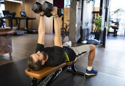 Bearded adult sportsman lying on bench and exercising with dumbbells during weightlifting training in modern gym