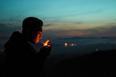 Side view of silhouette man smoking cigarette against sky at dusk