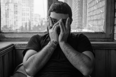 Portrait of young man with hands covering mouth sitting against window at home