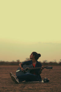 Young woman playing guitar while sitting on field against sky