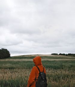 Rear view of person with backpack standing on grassy field against cloudy sky