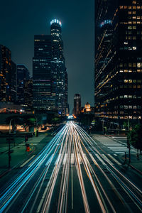 Light trails on road amidst buildings against sky at night