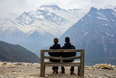 Rear view of couple sitting on bench against snowcapped mountains