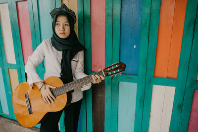 Full length of thoughtful woman playing guitar against wall