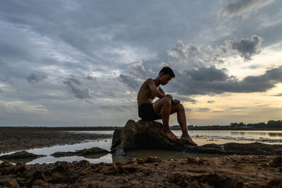 Shirtless boy sitting on rock against sky during sunset