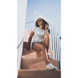 Low angle view of young woman wearing sunglasses while sitting on steps against sky