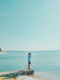 Side view of man photographing sea while standing on retaining wall against sky