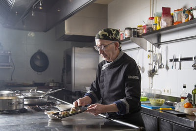 Mature male cook in uniform showing savory snack with fish and green guacamole served on plate in kitchen of restaurant