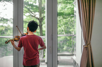 Rear view of boy playing violin by window