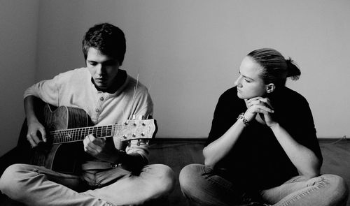 Young man playing guitar by friend on hardwood floor at home