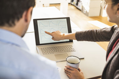 Businesswoman with coffee cup explaining diagram on laptop to businessman while sitting at office