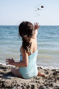 Rear view of girl throwing sand in sea while sitting at beach against clear sky