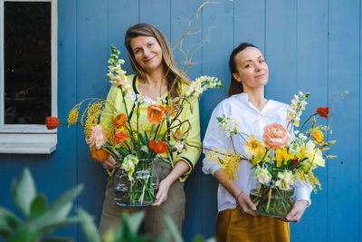 Two young women with beautiful bouquets of peonies in glass vases against the blue background