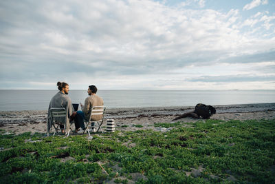 Male friends discussing over laptop while sitting on chairs at beach against sky