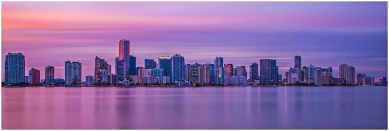 Sea by modern buildings against sky during sunset