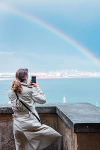 Rear view of woman photographing rainbow on sea