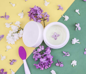 Face cream, lilac flowers and a brush for masks lie on a yellow-green background, close-up top view.