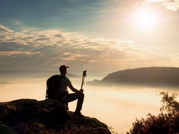 Man hiker with trekking poles and backpack on rock. old  bushes grows in rock, misty spring day.