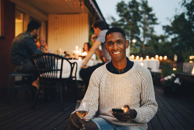 Portrait of smiling young man holding mobile phone while friends in background during dinner party