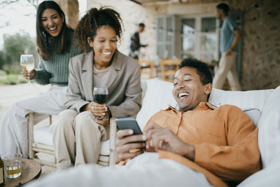 Cheerful man lying on sofa sharing smart phone with friends at backyard