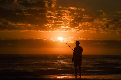 Silhouette man fishing at beach against sky during sunset