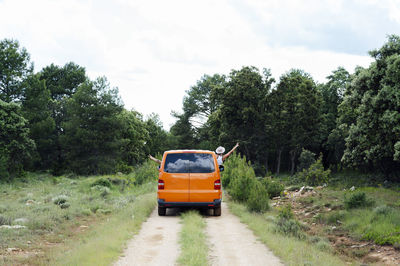 Carefree anonymous travelers with outstretched arms driving orange van along road in woods during summer trip