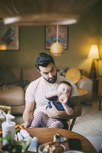 Baby girl with bottle sitting on father's lap in living room