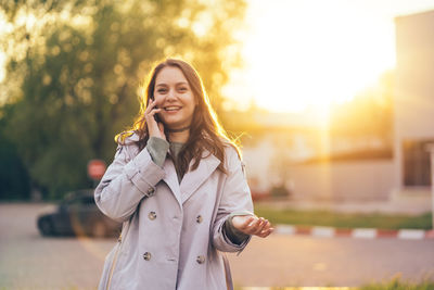 Smiling beautiful girl with long hair in a grey trench coat using smartphone in the spring 