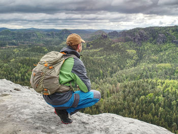 The body of man with a backpack and running shoes stands on top of a rock against rocky valley