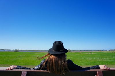 Rear view of woman wearing hat sitting on bench at field against clear blue sky during sunny day