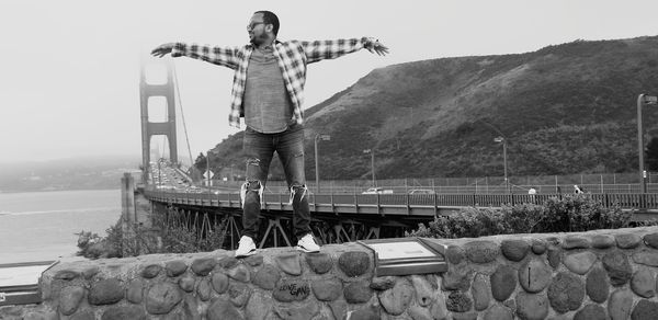 Man with arms outstretched standing on wall against sky