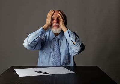 Senior businessman sitting with head in hand on table against gray background