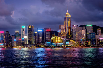 Illuminated city by river against cloudy sky at night