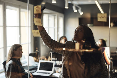 Female entrepreneur sticking adhesive note on glass wall during meeting in office