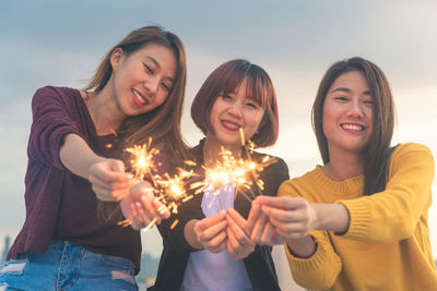 Portrait of smiling young women holding illuminated sparklers against sky