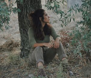 Full length of young woman sitting by tree on field
