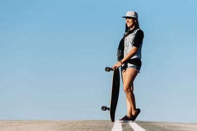 Full length of woman holding skateboard while standing on road against clear blue sky