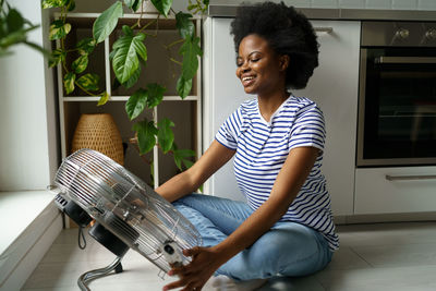 Happy smiling young black woman enjoying fresh air while cooling by electric fan at home
