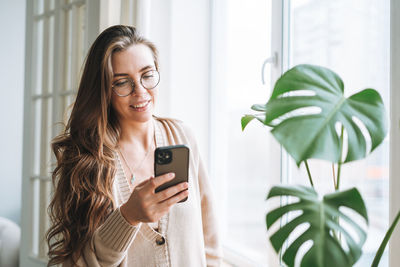 Young beautiful woman with brunette long hair in cozy knitted cardigan using mobile phone at home