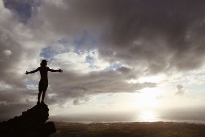 Silhouette man with arms outstretched standing on rock against sky during sunset