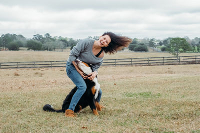 Cheerful owner and big bernese mountain dog have fun on field against village landscape.