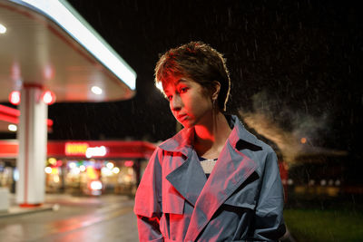 Upset lonely young millennial woman standing near petrol station shop in red neon light at night