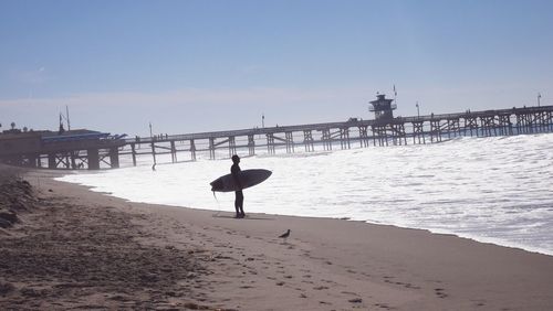 Man with surfboard standing at sea shore by pier against sky