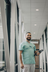 Smiling male healthcare worker holding file while standing in corridor of hospital