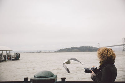 Side view of woman photographing bird while standing at pier over sea