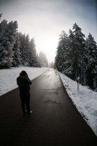 Rear view of man standing on road amidst snow covered pine trees during winter