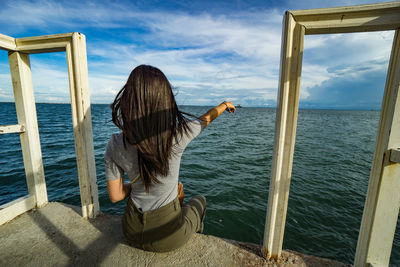 Rear view of woman sitting by sea against sky