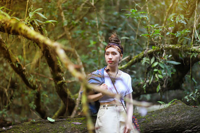 Thoughtful young woman standing by log against trees in forest