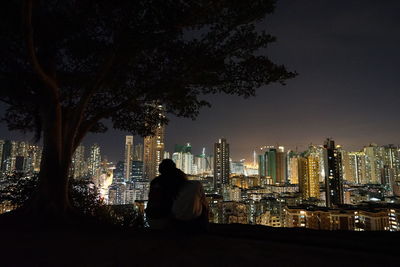 Rear view of man sitting against illuminated buildings at night