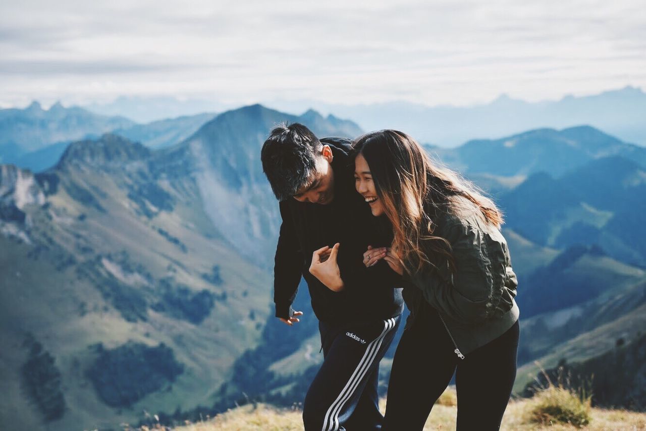 two people, mountain, love, togetherness, young women, real people, nature, heterosexual couple, leisure activity, young adult, standing, romance, bonding, casual clothing, mountain range, embracing, lifestyles, happiness, adventure, beauty in nature, women, young men, couple - relationship, men, outdoors, day, scenics, smiling, sky, friendship, people
